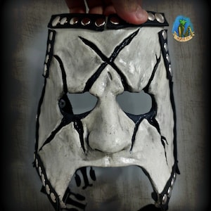 Puede incluir: Una máscara de cuero blanco con detalles pintados en negro, incluyendo una X en la frente y líneas alrededor de los ojos y la nariz. La máscara tiene una correa de cuero con tachuelas alrededor de la parte superior e inferior.