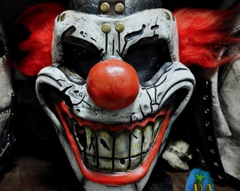 Puede incluir: Una máscara de payaso blanca y negra con una nariz roja y una amplia sonrisa con dientes. La máscara tiene una correa de cuero marrón con remaches metálicos y pelo rojo.
