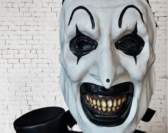 May include: A white clown mask with black accents, including eye shapes, eyebrows, and a wide, menacing grin revealing yellowed teeth. The mask has a black top hat and a black and white patterned head covering. The mask is designed to be scary.