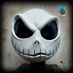 Puede incluir: Una máscara de cerámica blanca de Jack Skellington con ojos negros y una boca cosida. La máscara está sobre un soporte negro.