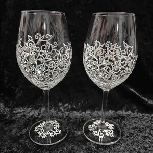 May include: Two clear glass wine glasses with a white lace design painted on the bottom half of each glass.