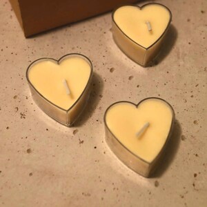 May include: Three heart-shaped tea light candles with wicks in clear glass holders. The candles are a pale yellow color and are arranged on a light brown surface.