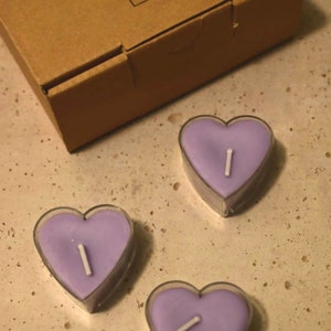 May include: Three purple heart-shaped candles with white wicks. The candles are in clear containers and are sitting on a light brown surface.
