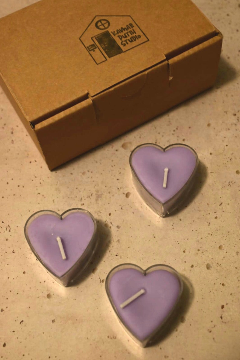 May include: Three purple heart-shaped candles with white wicks. The candles are in clear containers and are sitting on a light brown surface.