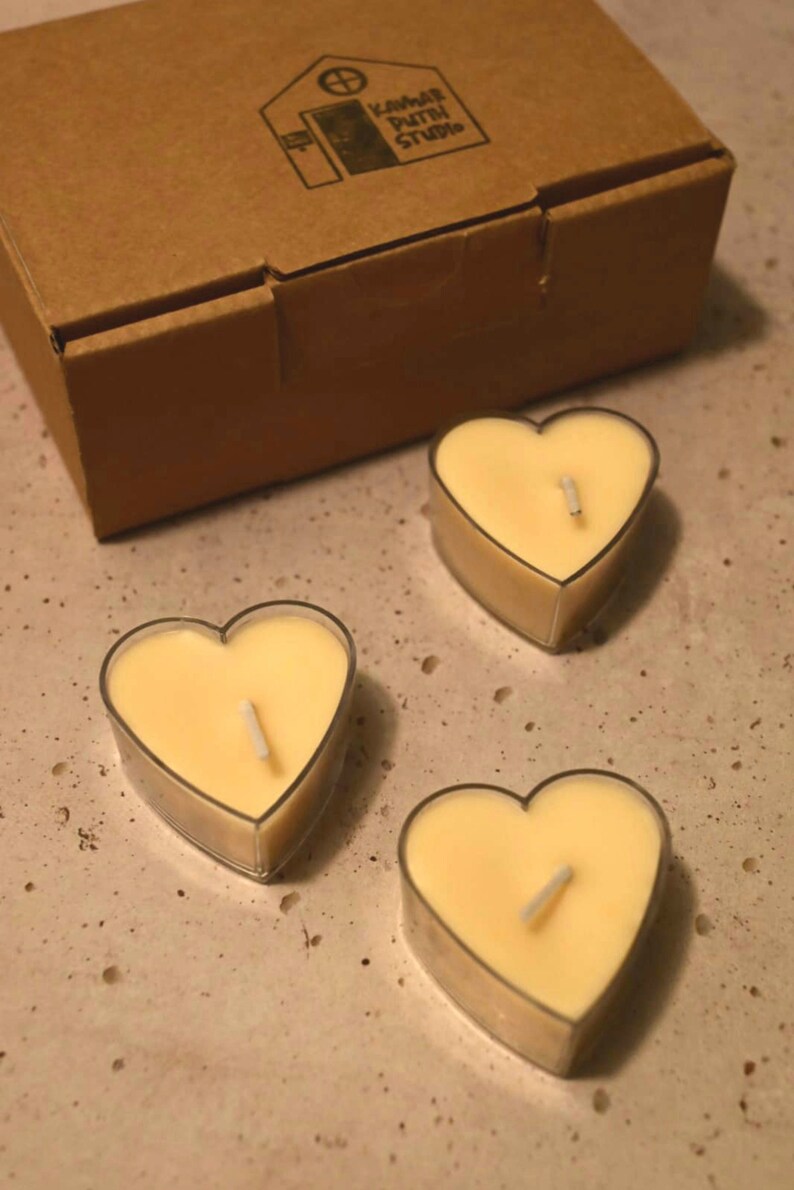 May include: Three heart-shaped tea light candles with wicks in clear glass holders. The candles are a pale yellow color and are arranged on a light brown surface.