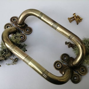 May include: Two antique brass door handles with a curved design and decorative detailing. The handles are accompanied by a small pile of brass screws. The handles are placed on a white surface with some moss.