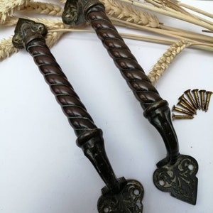 May include: A pair of antique door handles with a dark brown, twisted design. The handles have decorative, ornate ends and are accompanied by a small pile of brass screws. Wheat stalks are in the background.