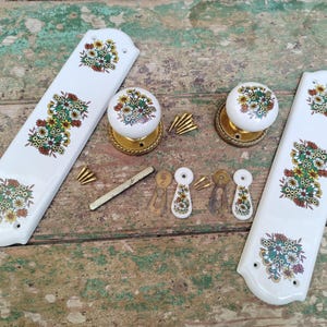 May include: A set of white ceramic door furniture with floral patterns in green, yellow, and red. The set includes two long plates, two round doorknobs with gold bases, and small decorative pieces.