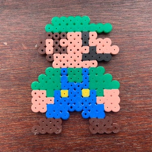 Op de afbeelding: Een pixel art-personage gemaakt van kleurrijke kralen. Het personage is groen, bruin, blauw en geel. Het personage draagt ​​een groene hoed en een overall.