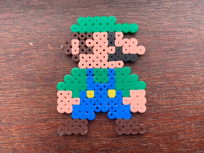 Op de afbeelding: Een pixel art-personage gemaakt van kleurrijke kralen. Het personage is groen, bruin, blauw en geel. Het personage draagt ​​een groene hoed en een overall.