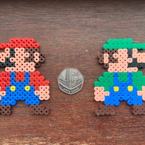 Op de afbeelding: Twee pixel art figuren van Mario en Luigi gemaakt met kleurrijke kralen. Mario draagt een rood shirt, een blauwe overall en een rode hoed. Luigi draagt een groen shirt, een blauwe overall en een groene hoed. Beide figuren hebben bruine schoenen.