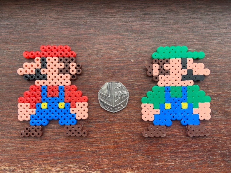 Op de afbeelding: Twee pixel art figuren van Mario en Luigi gemaakt met kleurrijke kralen. Mario draagt een rood shirt, een blauwe overall en een rode hoed. Luigi draagt een groen shirt, een blauwe overall en een groene hoed. Beide figuren hebben bruine schoenen.