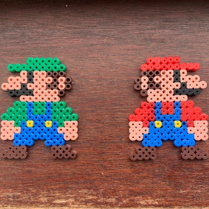 Op de afbeelding: Twee pixel art figuren van Mario en Luigi gemaakt met kleurrijke kralen. Mario draagt ​​een rode hoed en een overall, terwijl Luigi een groene hoed en een overall draagt.