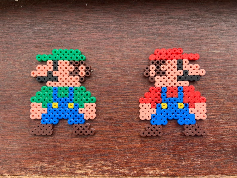 Op de afbeelding: Twee pixel art figuren van Mario en Luigi gemaakt met kleurrijke kralen. Mario draagt ​​een rode hoed en een overall, terwijl Luigi een groene hoed en een overall draagt.