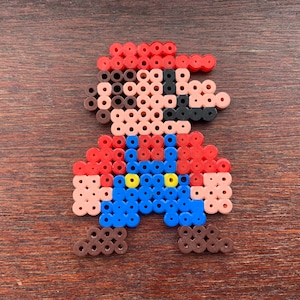 Op de afbeelding: Een pixel art-figuur gemaakt van rode, blauwe, bruine en gele kralen. Het figuur draagt een rode hoed en een overall.