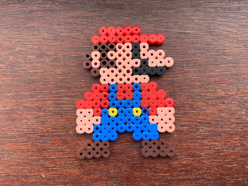 Op de afbeelding: Een pixel art-figuur gemaakt van rode, blauwe, bruine en gele kralen. Het figuur draagt een rode hoed en een overall.