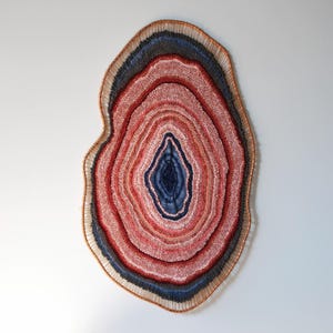 Könnte beinhalten: Ein großer, abstrakter Wandbehang mit einem geschichteten Design. Er zeigt konzentrische Ringe in Rot-, Blau- und Brauntönen, die einem Querschnitt durch eine Geode ähneln. Das Kunstwerk ist an einer weißen Wand befestigt und erzeugt einen auffälligen Kontrast.