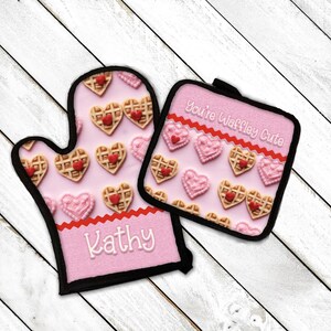 May include: Pink oven mitt and pot holder set with a waffle heart pattern and the text "You're Waffle-y Cute". The oven mitt has the name "Kathy" embroidered on it.