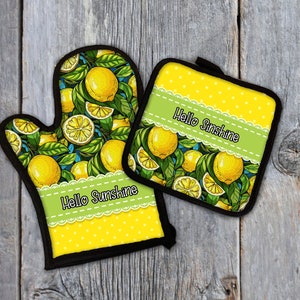 May include: A set of two yellow and green lemon-themed oven mitts. The larger mitt has a yellow background with white polka dots and a green ribbon with the text "Hello Sunshine". The smaller mitt has a yellow background with white polka dots and the text "Hello Sunshine" on a green ribbon.