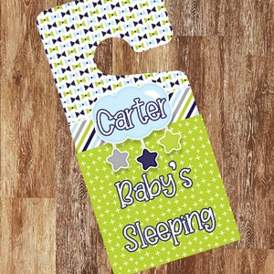 Puede incluir: Un colgador de puerta verde y azul con una nube blanca que dice "Carter" y el texto "Baby's Sleeping".
