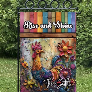 May include: A colorful garden flag with a rooster and flowers. The flag has a wooden background with the text "Rise and Shine" and "The Smiths".