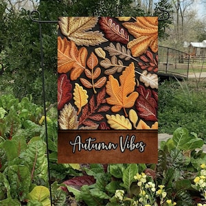 Könnte beinhalten: Eine Gartenflagge mit dunklem Hintergrund und einem Muster aus bunten Herbstblättern. Der Text "Autumn Vibes" ist unten auf der Flagge aufgedruckt.