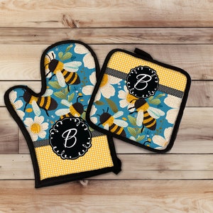 May include: A set of two yellow and blue kitchen pot holders with a bee and daisy pattern. The pot holders have a black border and a black circle with the letter "B" in the center.