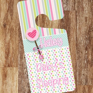 Pode incluir: Pendurador de porta listrado rosa e verde com fundo branco de bolinhas. O pendurador tem um recorte para uma maçaneta de porta e diz "Haley's Baby's Sleeping" com um coração rosa e um gráfico de chupeta.