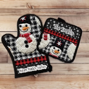Puede incluir: Un juego de dos manoplas para horno acolchadas en blanco y negro con detalles de cuadros rojos y blancos. Las manoplas tienen bordados de muñecos de nieve con ojos negros, narices naranjas y bufandas rojas. Una manopla tiene el texto "Warm Winter Wishes" y la otra tiene el texto "The Smiths".