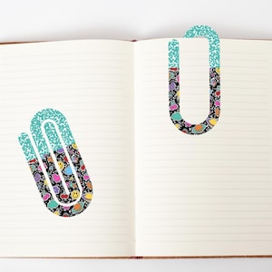 Puede incluir: Dos clips de marcador de página con estampado turquesa y negro. El patrón incluye imágenes de dibujos animados coloridos de nubes, estrellas, cerezas y caras sonrientes.