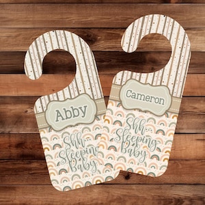 May include: Two wooden door hangers with a hook, featuring a striped top and a rainbow pattern. One reads "Abby" and the other "Cameron" in a decorative frame. The text "Shh Sleeping Baby" is also displayed.
