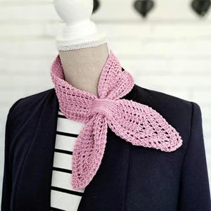 Op de afbeelding: Een lichtroze, open geweven gehaakte sjaal is om de nek van een mannequin gedrapeerd. De sjaal is in een knoop gelegd, met de uiteinden uitwaaierend. De mannequin draagt een marineblauwe blazer over een zwart-wit gestreept shirt.