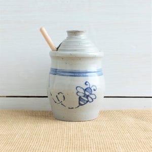 May include: A light gray speckled ceramic honey pot with a matching lid and a wooden dipper. The pot features a hand-painted blue bee motif on the front and a horizontal blue band around the upper section. The lid has a small hole for the dipper.