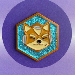 May include: A purple card with a white circle and two smaller circles at the bottom. A hexagonal pin with a blue glitter background and a gold outline features a cartoon fox with a white helmet and a blue visor. The fox is smiling and has a light brown body.