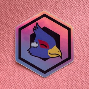 May include: A hexagonal sticker featuring a blue bird-like character with a yellow beak, red visor, and white eye covering. The character is set against a gradient background of pink, purple, and blue. The sticker has a black border.