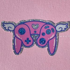 May include: A pink and blue video game controller with wings. The controller has a heart on the top and is covered in glitter.