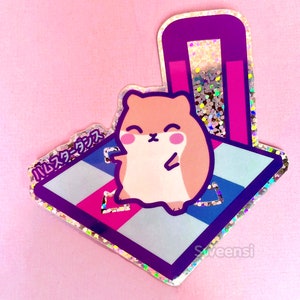 May include: A holographic sticker featuring a cartoon hamster wearing a pink and blue striped shirt, standing on a pink, blue and white chequered dance floor. The text "ハムスターダンス" is written in Japanese on the sticker.