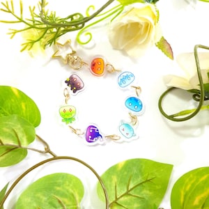 Puede incluir: Una pulsera de abalorios con abalorios de criaturas de dibujos animados de colores. Los abalorios están conectados por una cadena dorada y tienen un cierre en forma de estrella. El fondo incluye hojas verdes y flores blancas.