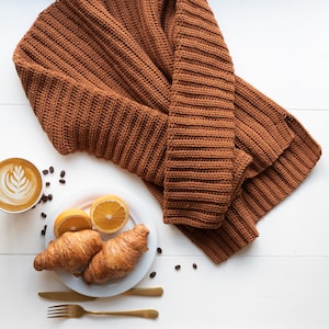 May include: A brown knitted jumper, two croissants, two orange slices, and a cup of coffee with latte art on a white surface.
