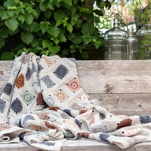 Puede incluir: Una manta de ganchillo con un patrón de cuadrados de la abuela multicolor en tonos de marrón, gris, rosa y verde. La manta está drapeada sobre un banco de madera.