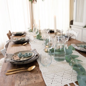 Può includere: Un tavolo in legno rustico apparecchiato per una cena con un runner da tavolo in crochet bianco, verde e posate dorate. Il tavolo è decorato con rami di eucalipto e candele.
