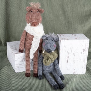 May include: Two crocheted stuffed animals, one brown with a white scarf and one gray with a green scarf, sitting on a white wooden box.