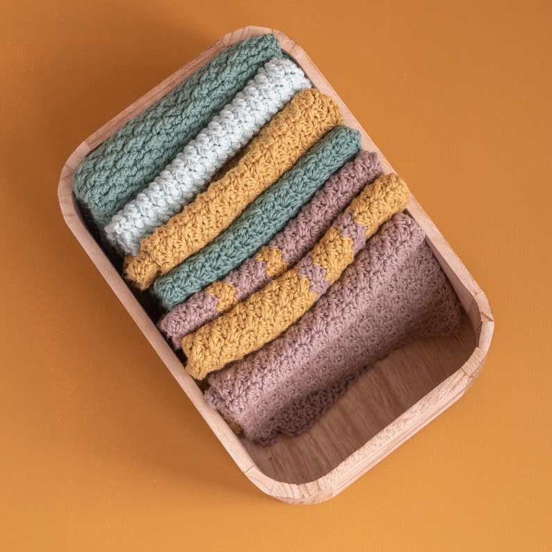 May include: A wooden box filled with a stack of crocheted dishcloths in various shades of green, yellow, and brown.