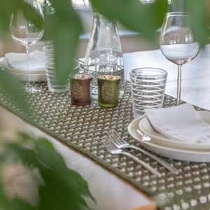 Può includere: Un runner da tavolo in crochet verde e bianco con due piccoli portacandele dorati e verdi, due bicchieri trasparenti e un tovagliolo bianco su un piatto bianco.