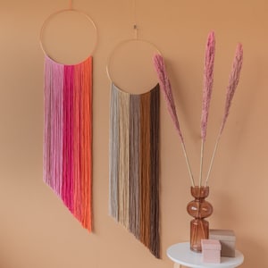 May include: Two macrame wall hangings with colorful yarn strands, one with pink, red, and orange strands and the other with beige, brown, and tan strands. The hangings are suspended from gold hoops and are displayed against a light brown wall.
