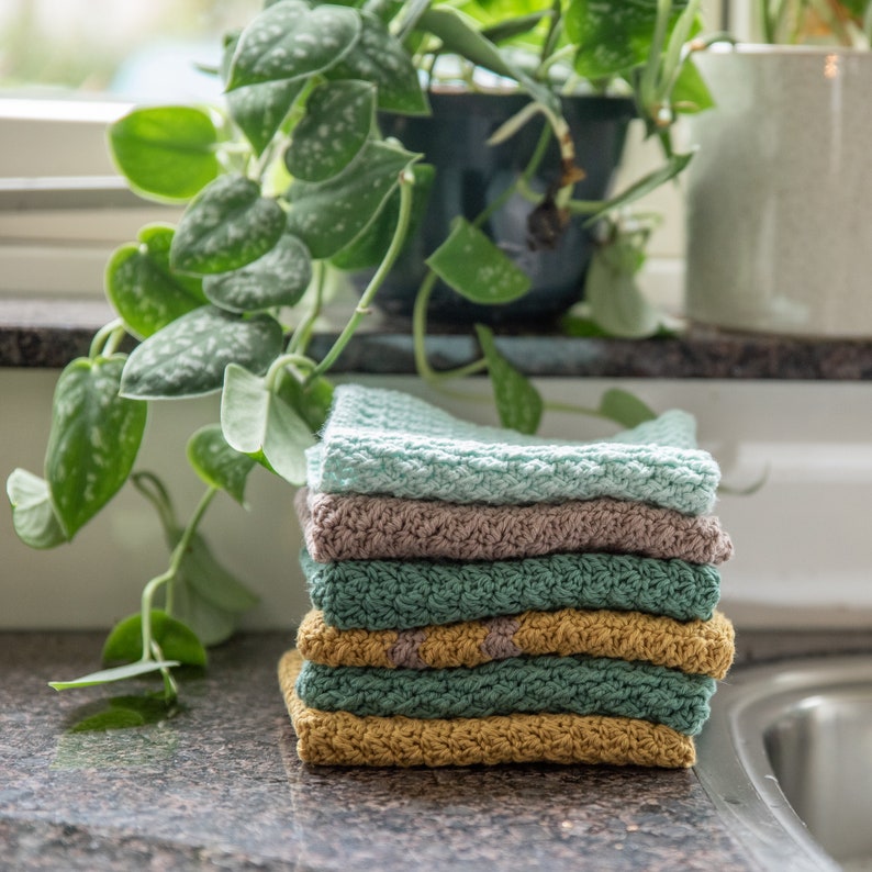 May include: A stack of six crocheted dishcloths in various shades of green, brown, and yellow. The dishcloths are folded and stacked on a countertop.