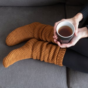 Puede incluir: Una persona con polainas de punto de color marrón se sienta en un sofá gris sosteniendo una taza negra de té.