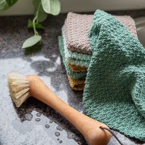 May include: A stack of four crocheted dishcloths in shades of green, brown, and yellow. The cloths are made with a textured stitch and are sitting on a gray countertop. A wooden brush with natural bristles is also in the image.