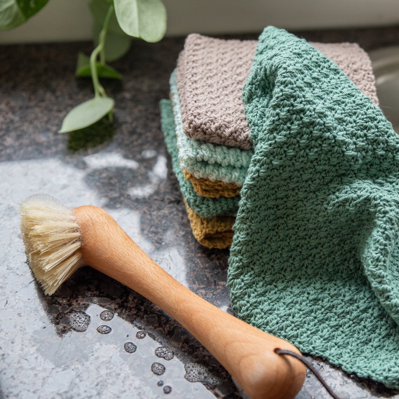 May include: A stack of four crocheted dishcloths in shades of green, brown, and yellow. The cloths are made with a textured stitch and are sitting on a gray countertop. A wooden brush with natural bristles is also in the image.