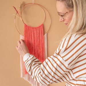 May include: A pink and coral yarn wall hanging with a copper hoop and fringe. The hanging is attached to a beige wall.